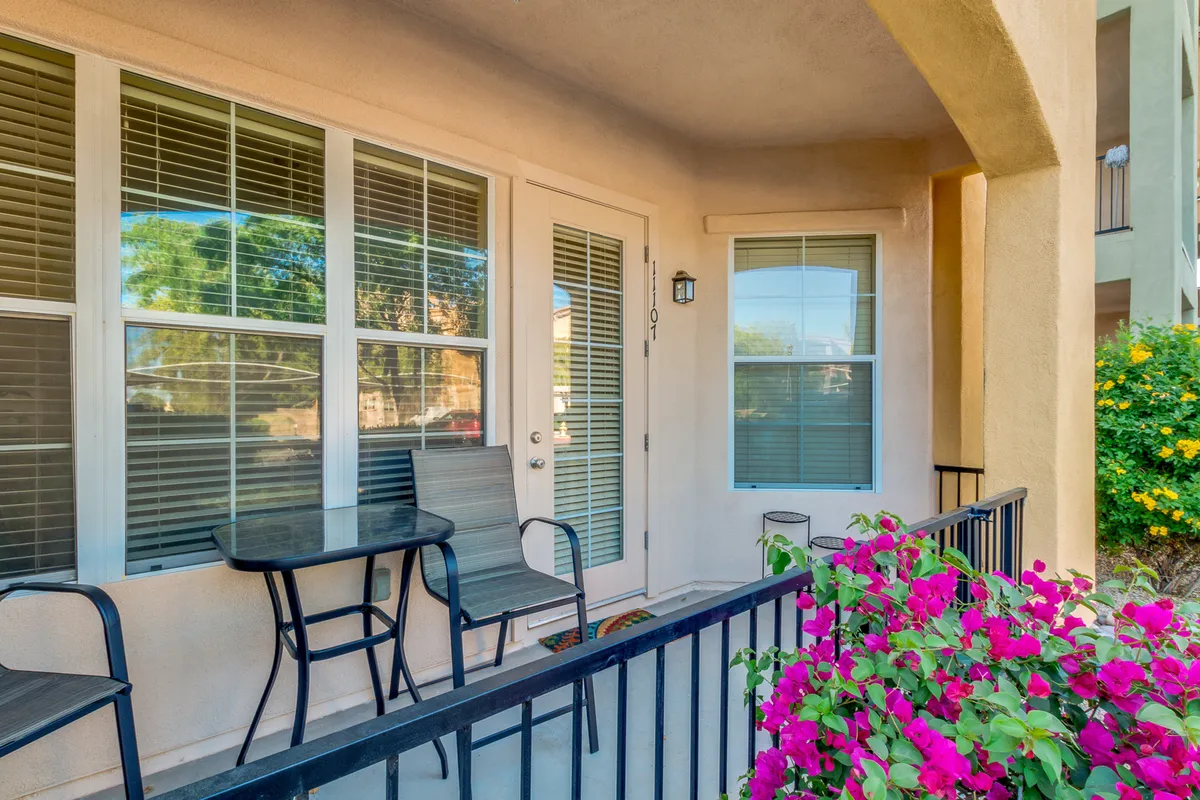 A cozy balcony setting featuring a table and chairs surrounded by flowering plants, adjacent to a doorway. The arrangement creates an inviting space for sipping morning coffee, enjoying the view, or relaxing amidst greenery.