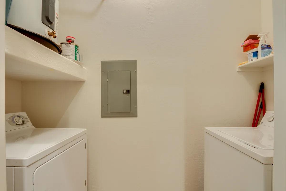 A practical laundry area is shown with a washing machine, dryer, organized shelves holding cleaning supplies, and a visible circuit breaker panel. The space is neat and functional, suited for everyday tasks.