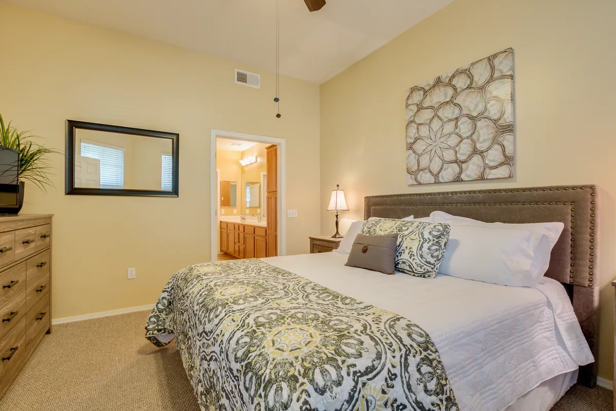 A serene bedroom featuring a king-sized bed with decorative bedding, complemented by framed wall art and a dresser with a mirror. Sunlight streams in, creating a calm and inviting atmosphere ideal for rest and relaxation.
