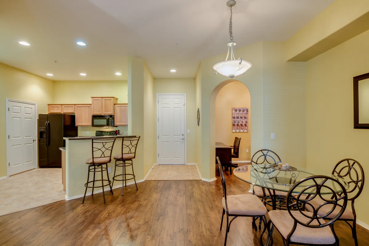 An inviting dining area with a glass table surrounded by chairs, adjacent to a kitchen counter with bar stools. An arched entrance connects to another room, emphasizing an open, flowing layout ideal for entertaining, family meals, or casual gatherings.