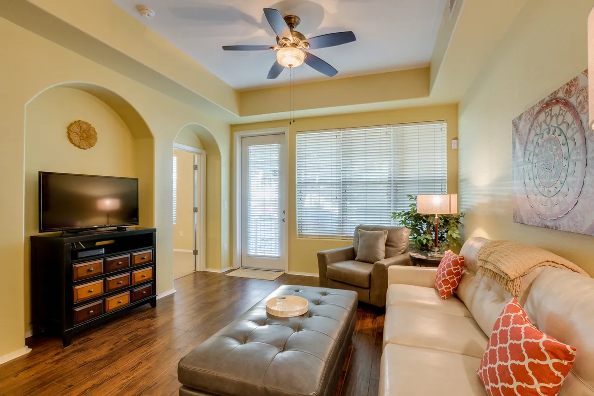 A cozy, inviting living room featuring a cream-colored sectional sofa, a dark wooden coffee table, a stylish television unit with decorative drawers, a large window with blinds that brings in natural light, and thoughtful wall art accents. The setup encourages relaxation, conversation, and media enjoyment.