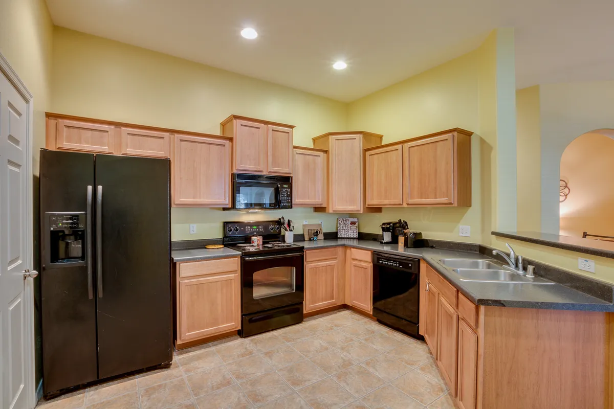 This image features a modern kitchen with a black refrigerator, wooden cabinetry, stainless steel appliances, and spacious countertops. The kitchen appears well-lit and is clearly designed for both cooking and entertaining, with room for multiple activities.