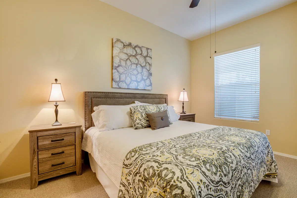 This image presents a cozy bedroom with a well-made bed flanked by nightstands and topped with elegant wall art. Sunlight streams through the window, illuminating the inviting space and suggesting a tranquil, restful environment ideal for extended stays.