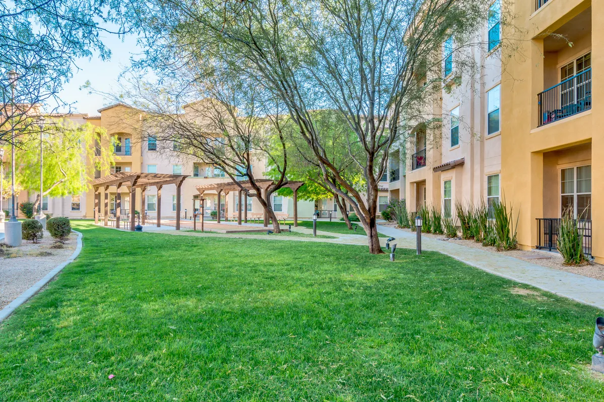 A broad exterior view highlighting green grass lawns, a walkway, mature trees, building facades, and a pergola. The setting invites a sense of community, ideal for picnics, group activities, and evening strolls.