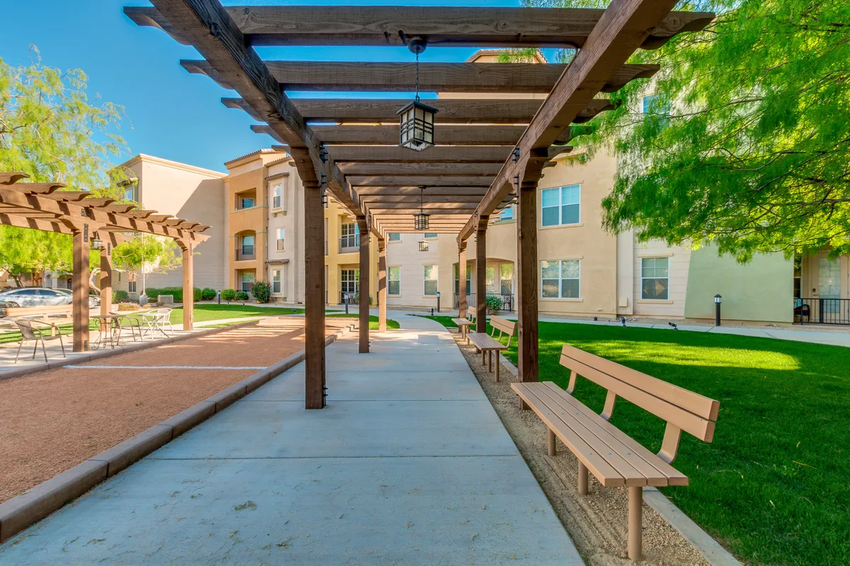 An exterior scene with a pergola structure over a landscaped pathway, benches for sitting, and lush, well-kept gardens. The tranquil mood is set by people walking, socializing, and relaxing in a communal space.