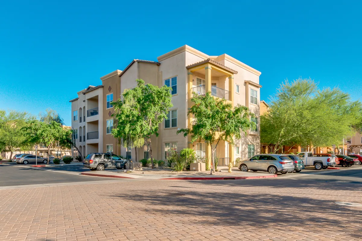 This exterior image shows the apartment building surrounded by lush greenery, with a visible parking area and open courtyard. The welcoming outdoor environment suggests opportunities for socializing, enjoying sunny days, and participating in community events.
